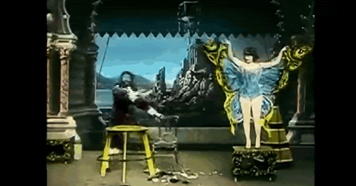 Video clip of a magician on a stage holding a chair and gesturing to a woman in a blue and yellow butterfly costume.