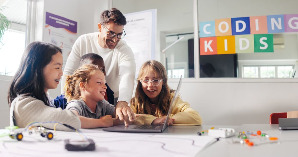 Male teacher guides elementary school students in a robot programming class. Kids learn to code using education software on a laptop and control robot vehicles in a practical computer science lesson